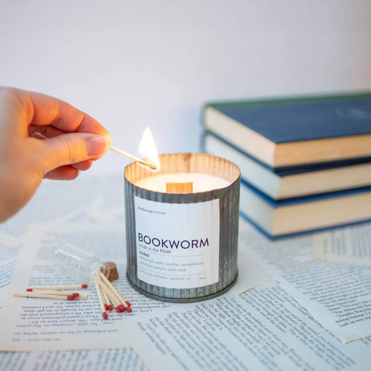 Person lighting a Bookworm candle surrounded by books and matchsticks on a scattered paper background.