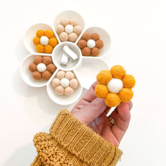 A hand holding a yellow felt flower with a round felt insert, surrounded by a selection of neutral and yellow felt flowers.