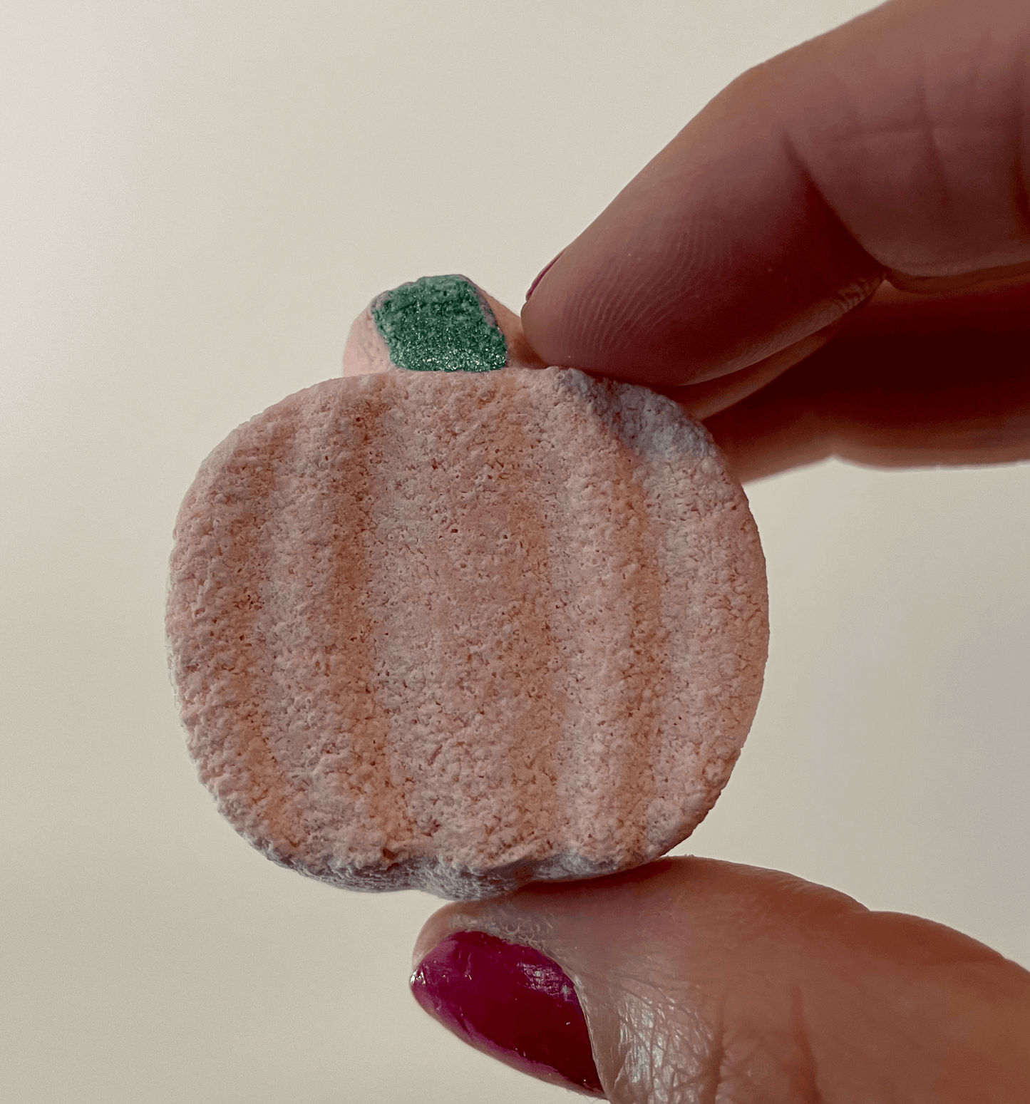 Natural mini pumpkin bath bomb, held up with fingers with pink nail polish.