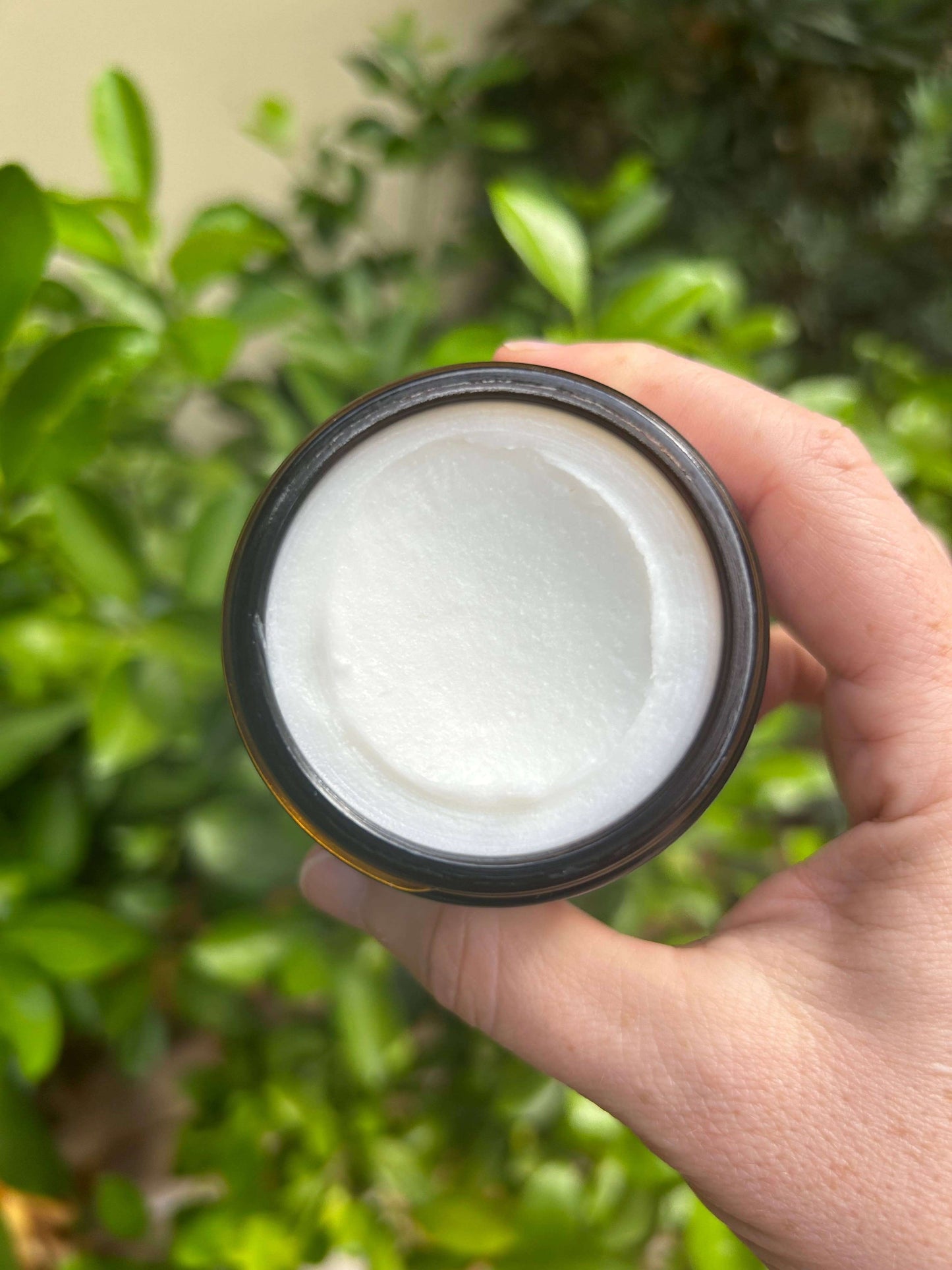 Hand holding a jar of Magnesium Sleep Butter, showcasing its creamy texture, surrounded by green foliage.