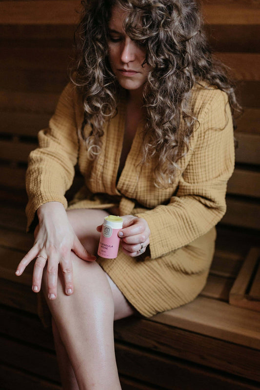 Woman applying calendula rose herbal salve from eco-friendly tube in a serene sauna setting.