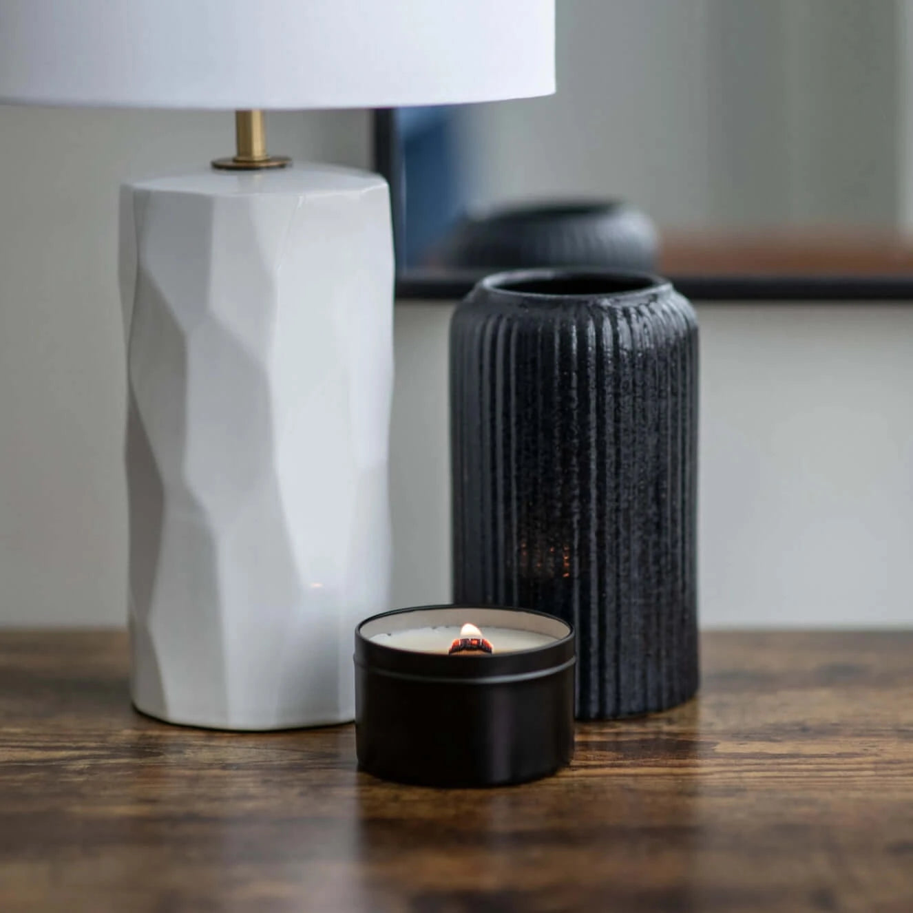 Black Travel candle with burning wood wick sitting in front of a white lamp and black vase.
