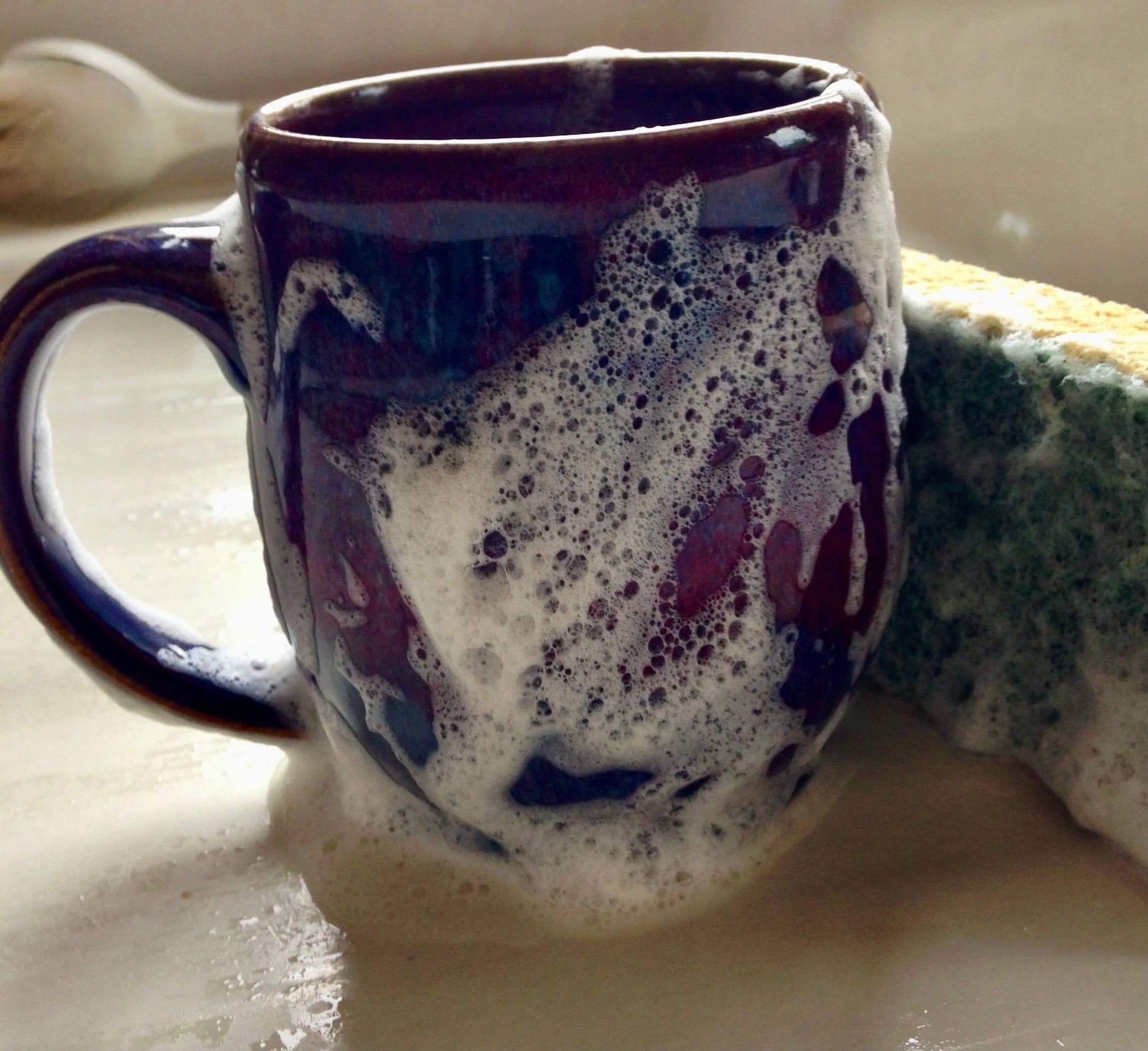 Foamy lavender liquid soap in a mug next to a green sponge, demonstrating effective cleaning power.