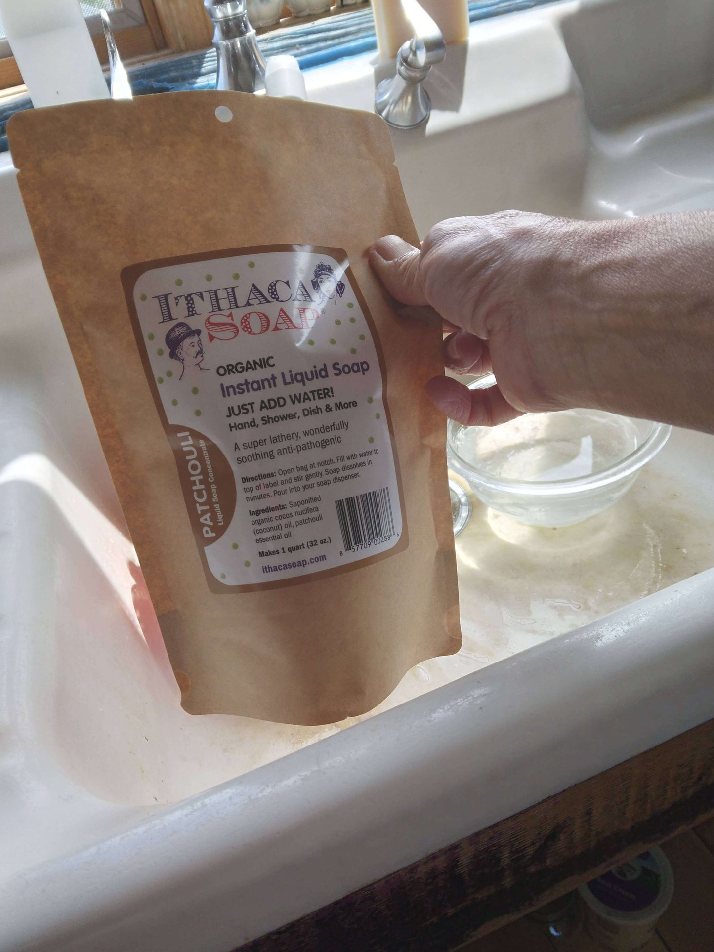 Person holding Ithaca Soap Instant Liquid Soap refill bag in a kitchen sink setting.