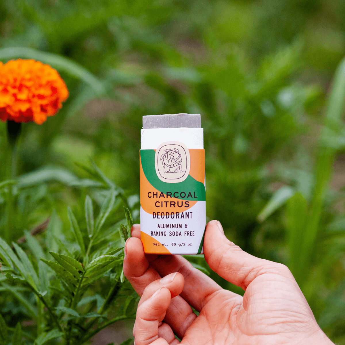 A hand holding a charcoal citrus deodorant stick outside in front of green plants and an orange flower.