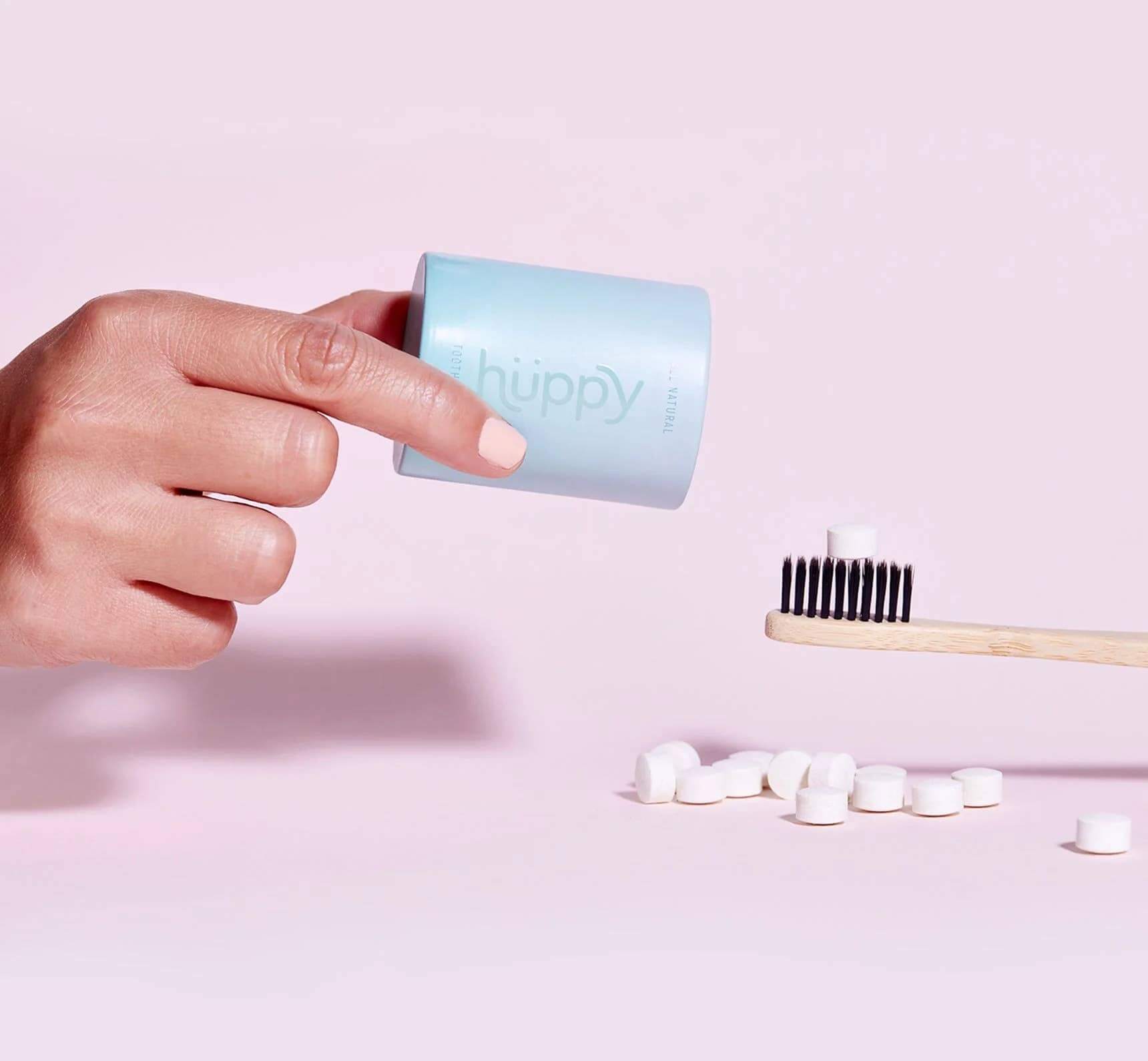 Hand holding Hüppy toothpaste tablet container above a bamboo toothbrush with white tablets on a pastel background.