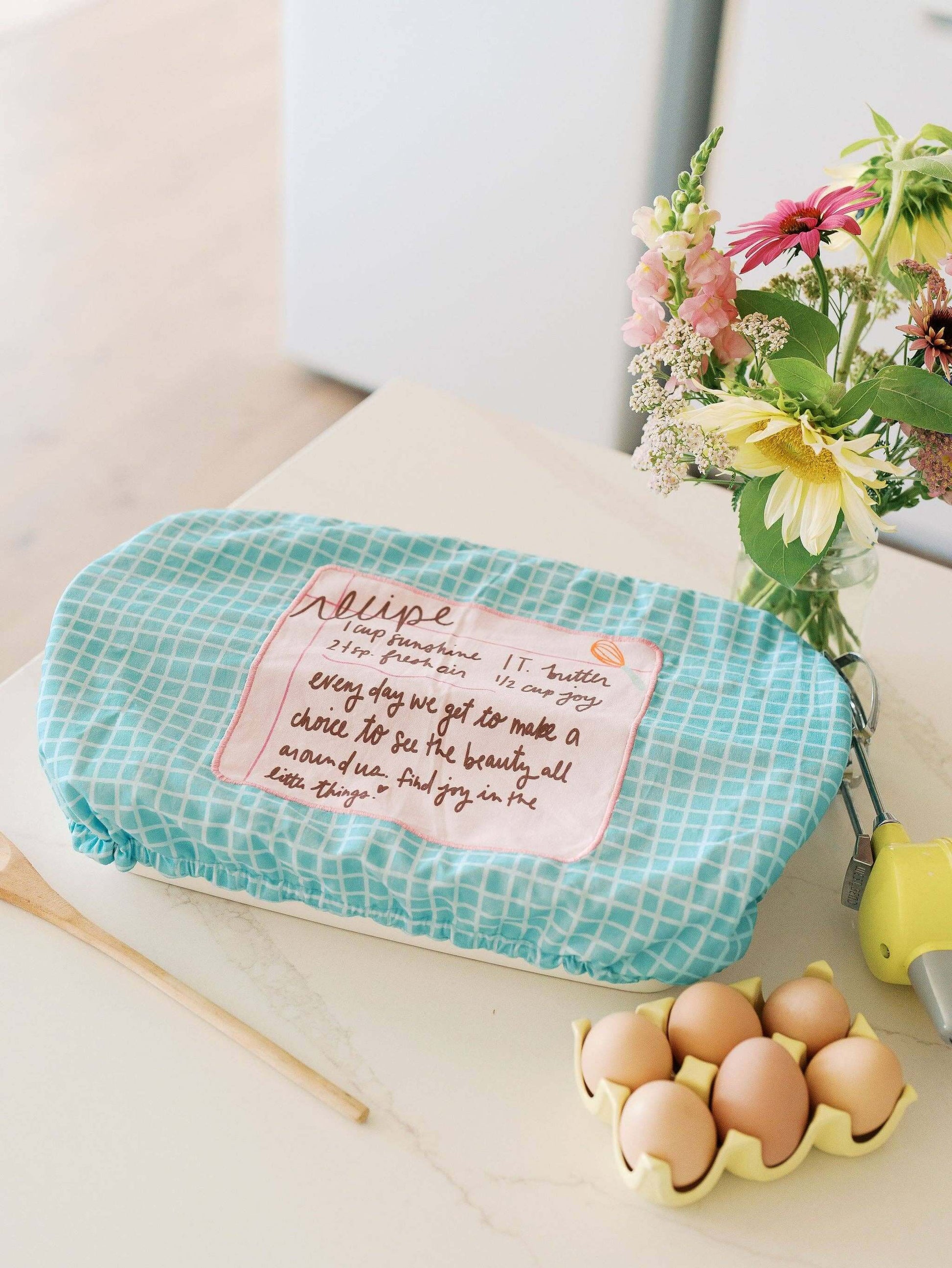Recipe for a Happy Heart 10x14 Baking Dish Cover | Baking with floral arrangement and eggs on a countertop.