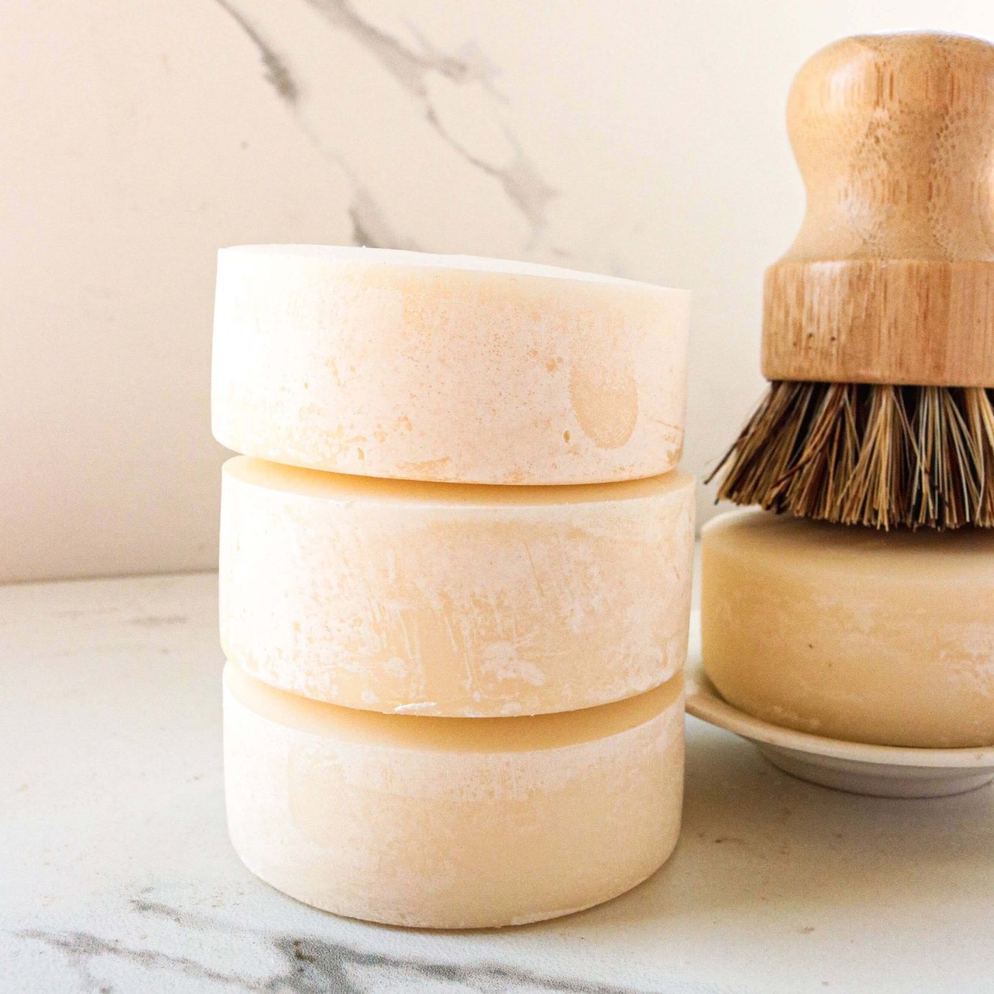 Three stacked solid dish soap bars next to a wooden dish brush on a marble surface.