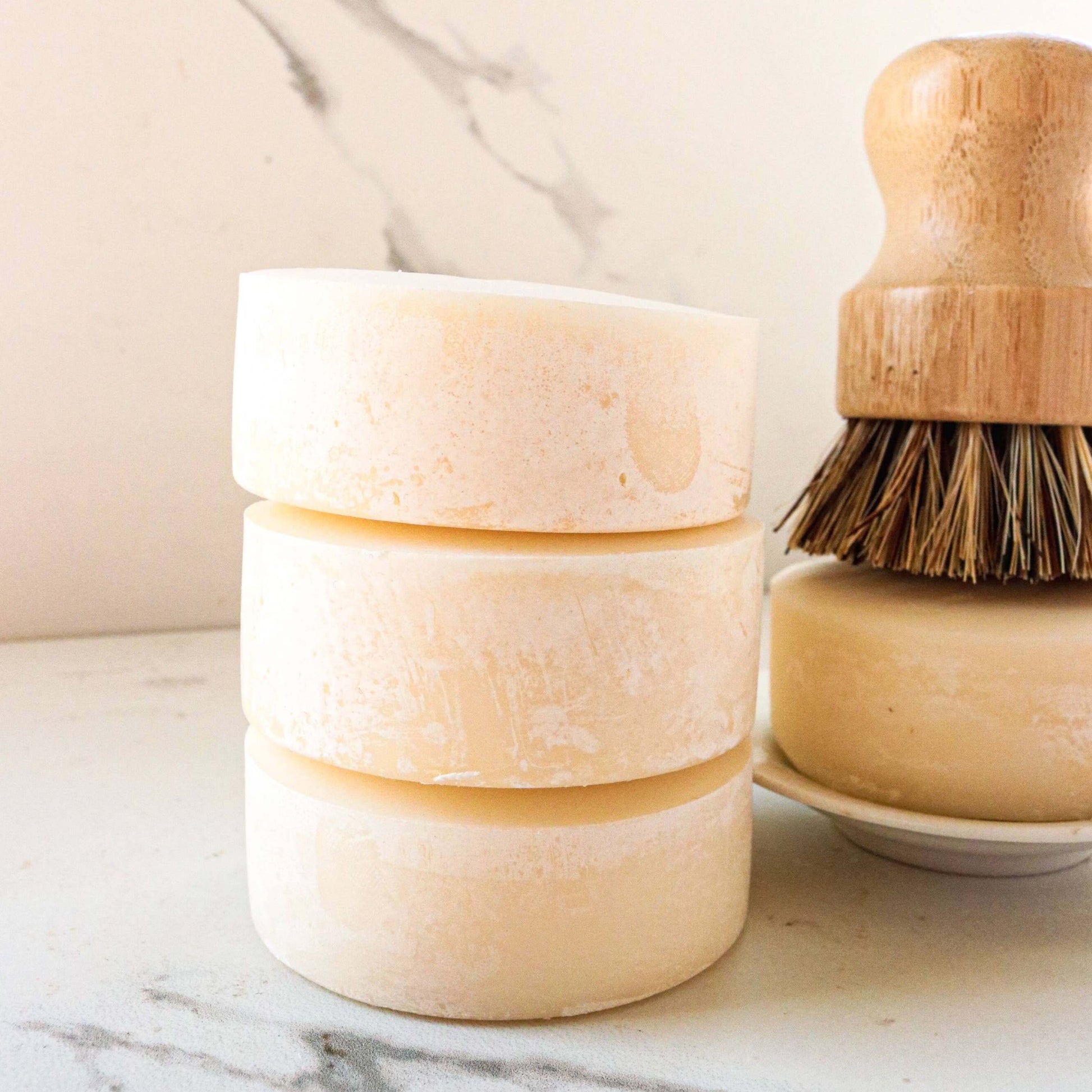 Three stacked solid dish soap bars next to a wooden dish brush on a marble surface.