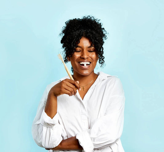 A lady in a white shirt holding up a bamboo toothbrush.  She is smiling with a toothpaste tablet in between her front teeth.
