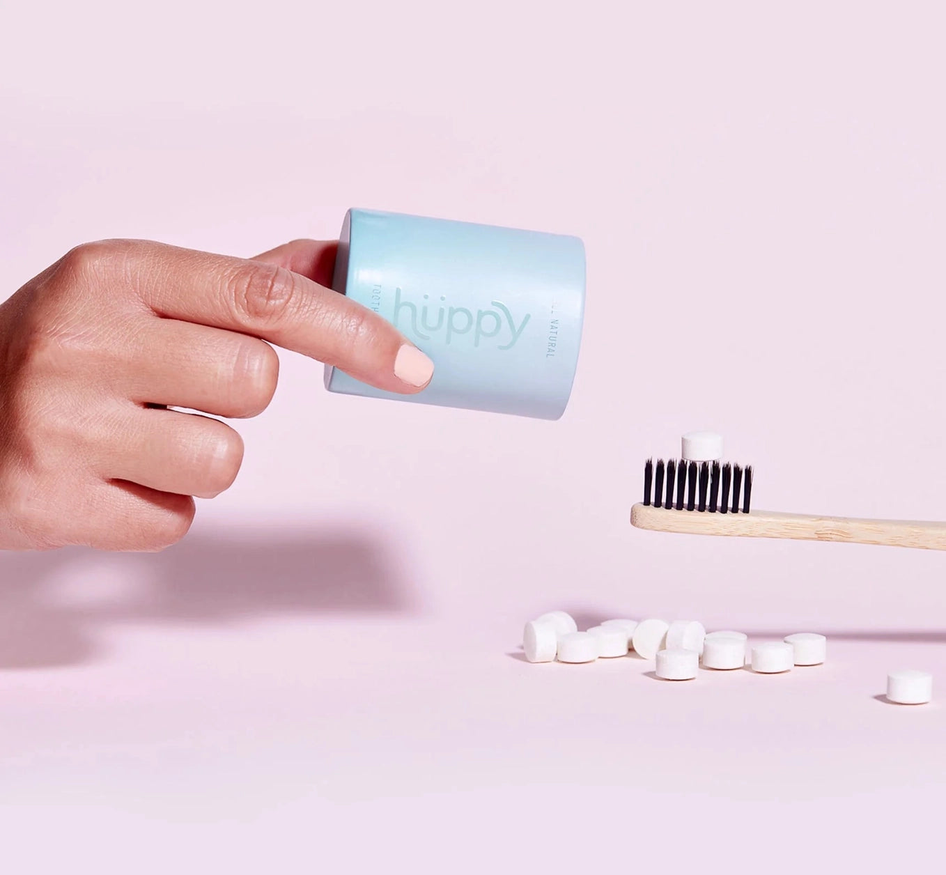A hand holding a Huppy toothpaste tablet container with toothpaste tablets on the table and a bamboo toothbrush with a tablet on it.