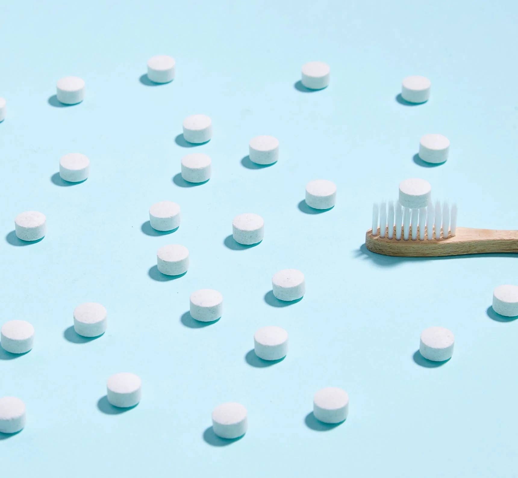 Toothpaste tablets spread around and a bamboo toothbrush with a toothpaste tablet on it with a light blue background.