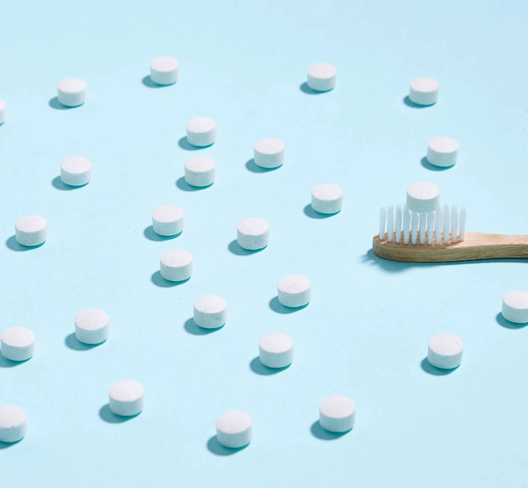 Toothpaste tablets spread around and a bamboo toothbrush with a toothpaste tablet on it with a light blue background.