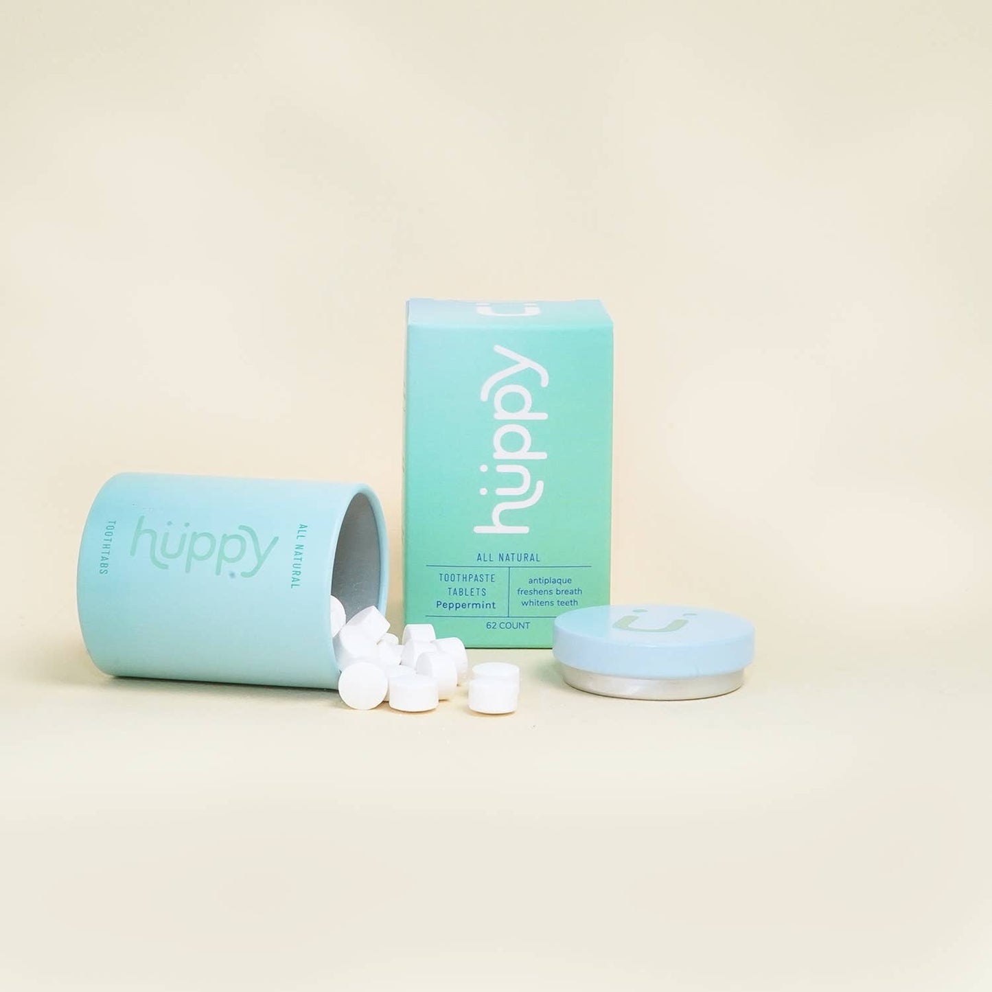 Huppy toothpaste tablets in a blue round metal container, laid on its side with tablets coming out.  The blue box and blue lid are sitting next to them.  