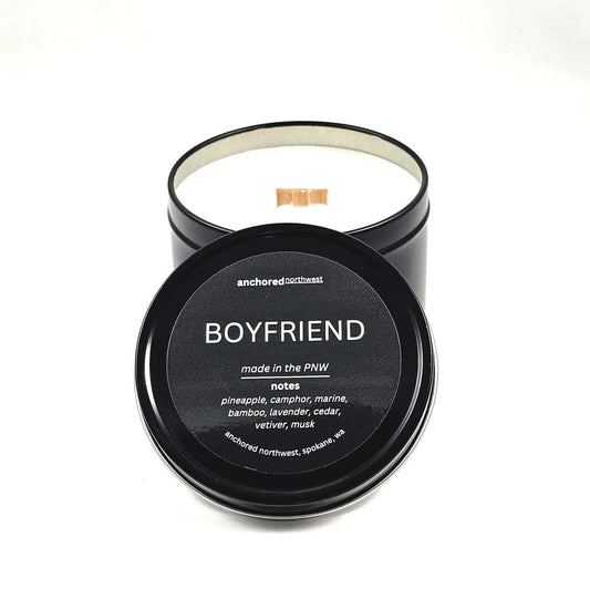 Non Toxic soy candle in a black tin with a wood wick.  Lid is sitting in front with the anchored northwest label and boyfriend scent.