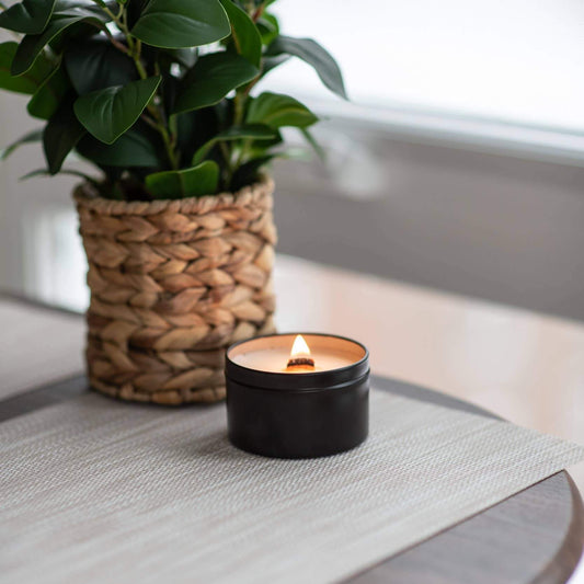 Black travel candle with a lit wood wick sitting on a round side table with a plant behind it.
